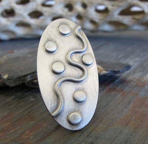 Oval silver ring with dots and wavy line on rock with driftwood